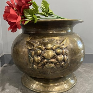 May include: An antique brass planter with a detailed, embossed lion-like face. The planter has a wide, rounded body and a flared rim, and is filled with red roses. The planter sits on a pedestal base.