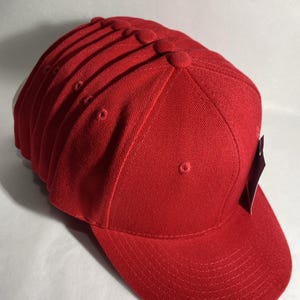 May include: A stack of red baseball caps. Each cap has a curved brim, a rounded top, and a small button on top. The caps are made of a textured fabric and appear to be new, with tags attached.
