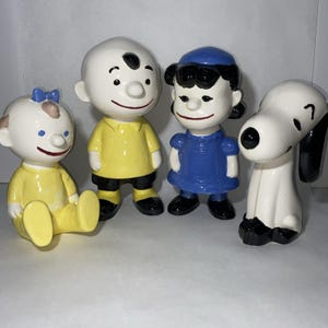 May include: Four ceramic figurines of Peanuts characters. The figures include Charlie Brown in a yellow shirt, Lucy in a blue dress, a baby in yellow, and Snoopy. The figurines are painted with black, white, yellow, and blue.