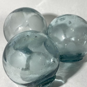 Puede incluir: Tres boyas redondas de vidrio transparente en diferentes tonos de azul-verdoso. Las esferas tienen una superficie ligeramente texturizada con burbujas e imperfecciones visibles. Las boyas están dispuestas muy juntas, sugiriendo un tema náutico o decorativo.