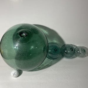 Pode incluir: Um grande flutuador de vidro verde com múltiplas esferas conectadas. O flutuador tem uma forma arredondada com uma característica proeminente em forma de olho. Uma bola de golfe é colocada por baixo para escala. O vidro tem uma superfície ligeiramente texturizada.