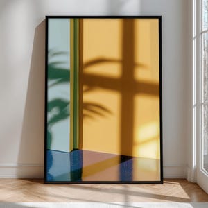 May include: Framed art print depicting an abstract interior scene. The artwork features a yellow wall with a plant shadow, a blue and pink floor, and a green and yellow vertical element. The print is enclosed in a black frame.