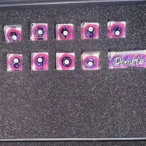 May include: A set of eleven square resin keycaps featuring a swirling pink and purple galaxy pattern with a central circular eye design. Each keycap displays a black number or symbol, and one rectangular keycap features the word Dimple in script.