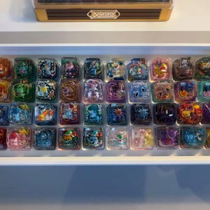 May include: A collection of forty square resin keycaps arranged in a white rectangular tray. Each individual keycap features a unique, colourful miniature character figure encased within clear, glossy resin, showcasing various detailed designs.