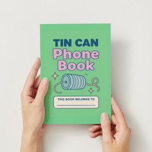 Puede incluir: Un cuaderno verde titulado "Tin Can Phone Book" con una ilustración retro de un teléfono de lata. La portada también incluye el texto "This Book Belongs To" con una línea en blanco. El cuaderno está sostenido por dos manos.
