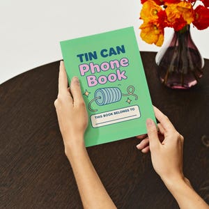 May include: A green "Tin Can Phone Book" notebook with the text in blue and pink. The notebook has a drawing of a spool of thread and the words "This book belongs to" at the bottom. The notebook is held by a person.