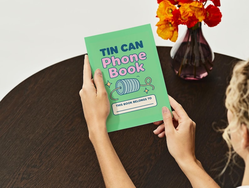 May include: A green "Tin Can Phone Book" notebook with the text in blue and pink. The notebook has a drawing of a spool of thread and the words "This book belongs to" at the bottom. The notebook is held by a person.