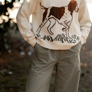 May include: A cream-colored long-sleeve knit sweater featuring a brown and white dog graphic on the front, paired with light green trousers that have a white grid pattern. The sweater has a ribbed crew neckline, cuffs, and a ribbed hem.