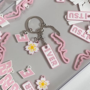 Puede incluir: Un llavero plateado con múltiples colgantes rosas y blancos. Los colgantes incluyen una flor de cerezo, la palabra "IVER" y una forma de "H" estilizada. También son visibles colgantes rosas adicionales con la palabra "ITSU".