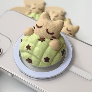 May include: A beige cat figurine with closed eyes resting inside a light green quilted sleeping bag decorated with small brown stars. The cat figure is attached to a circular white magnetic phone grip mount resting on a clear smartphone case.