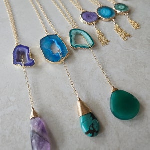 May include: A collection of gold necklaces with various gemstone pendants, including amethyst, turquoise, and green agate. The necklaces feature delicate chains and unique gemstone shapes.