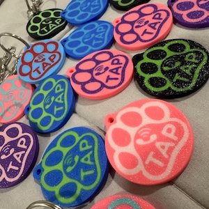 Può includere: Una collezione di portachiavi ovali colorati con un design a zampa e la parola "TAP" al centro. I portachiavi sono in vari colori, tra cui rosa, blu, verde e viola, con portachiavi argentati.