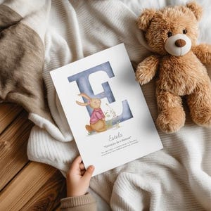 May include: A print featuring a blue letter "E" with a watercolor effect, a cartoon bunny wearing a pink sweater, and the name "Estela". A brown teddy bear is in the background.