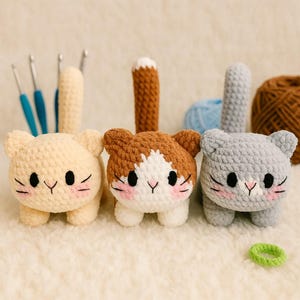 May include: Three handmade crochet cat toys in cream, brown and white, and gray. Each cat has black eyes, pink cheeks, and whiskers. Crochet hooks, yarn, and a green hair tie are in the background.
