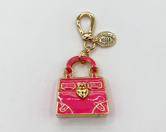 Puede incluir: Un charm de bolso de metal dorado con acabado de esmalte rosa. El charm presenta un diseño de bolso pequeño y detallado con un cierre en forma de corazón. Una pequeña etiqueta redonda dice "LIMITED EDITION 2014".