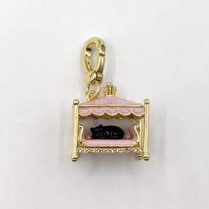 May include: A gold-coloured charm depicting a pink bed with a black animal figure inside. The bed is topped with a crown and decorated with rhinestones. The charm has a lobster clasp for attachment.