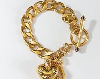 May include: Gold-coloured chain bracelet with a heart charm and a 'J' charm. The heart charm is engraved with "Juicy Couture". The bracelet features a toggle clasp with crown details.