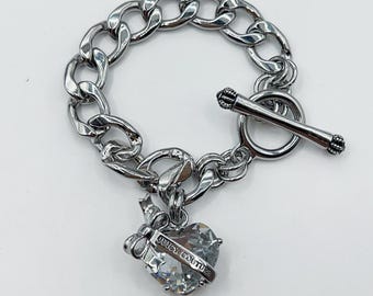 May include: Silver-toned chain bracelet with a toggle clasp. A heart-shaped charm with a clear crystal center and the words "Juicy Couture" is suspended from the chain. A small bow is attached to the charm.