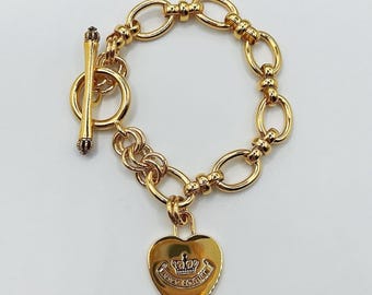 May include: Gold-toned bracelet featuring a toggle clasp and a heart-shaped charm. The charm is engraved with a crown and the words "Juicy Couture". The bracelet has an oval link chain design.