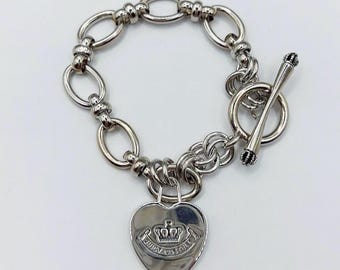 May include: Silver-toned bracelet featuring a heart-shaped charm with a crown design and the words "Juicy Couture". The bracelet has a toggle clasp and a chain of oval links.