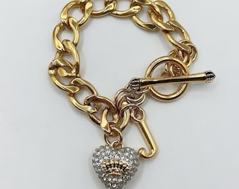 May include: Gold-tone chain bracelet with a toggle clasp. The bracelet features a heart-shaped charm encrusted with clear crystals and a gold crown design. A gold 'J' charm hangs next to the heart.