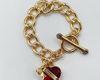 May include: Gold-tone chain bracelet with a toggle clasp. A red heart-shaped charm with the words "Juicy Couture" is attached. The charm is adorned with a gold bow. The bracelet is on a white background.