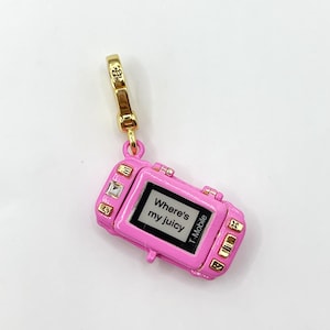 Può includere: Un ciondolo rosa a forma di telefono a conchiglia con dettagli color oro. Lo schermo mostra il testo "Where's my juicy" e il logo T-Mobile. Il ciondolo ha una chiusura color oro.