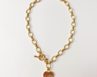 May include: Gold-toned necklace with an oval link chain and a heart-shaped pendant. The pendant features a crown design and text. The necklace has a toggle clasp closure. The jewelry is displayed on a white surface.
