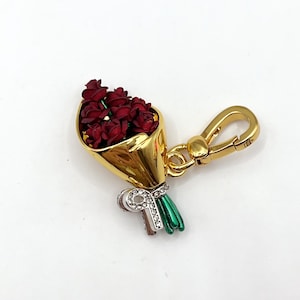 May include: A gold charm depicting a bouquet of red roses. The flowers are presented in a gold cone, tied with a green and silver bow. The charm includes a gold clasp for attachment to a keyring or bag.