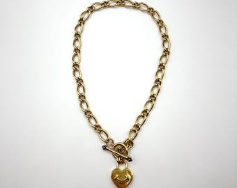 May include: Gold-toned necklace with a heart-shaped pendant. The pendant features a smiley face and is adorned with small, sparkling accents. The necklace has a toggle clasp and a chain link design.