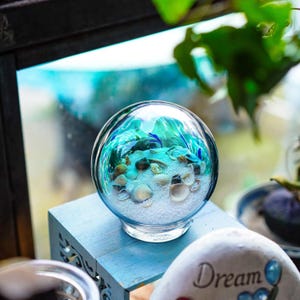 May include: A clear glass globe terrarium with a beach scene inside. The terrarium contains white sand, seashells, and blue and green decorative elements. The globe sits on a light blue wooden surface.