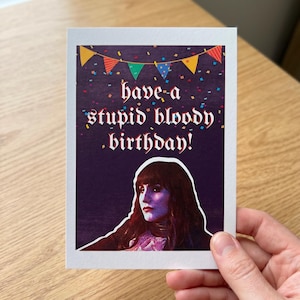 May include: A greeting card with the text "have-a stupid bloody birthday!" in white gothic font. The card features a portrait of a woman with long dark hair and a purple and blue face. Colorful confetti and bunting decorate the top.