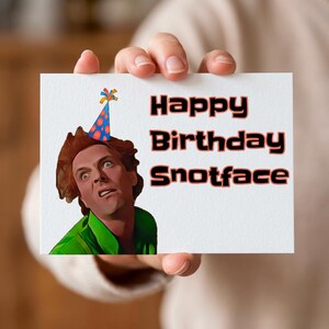Puede incluir: Una tarjeta de felicitaci&oacute;n blanca sostenida por una persona, con el texto "Happy Birthday Snotface" en negro y rojo. La tarjeta tiene una ilustraci&oacute;n de una persona con un gorro de fiesta y una camisa verde.