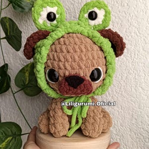 Puede incluir: Un perro pug de ganchillo hecho a mano con un sombrero de rana verde. El pug es marrón con una nariz marrón oscuro y ojos negros grandes. El sombrero de rana tiene detalles blancos y ojos negros grandes. El texto "@Liligurumi.Oficial" es visible.