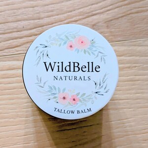 May include: A circular, white container with a black edge, displaying the text "WildBelle NATURALS TALLOW BALM." The lid is adorned with a floral wreath of pink roses and green foliage. The container rests on a wooden surface.