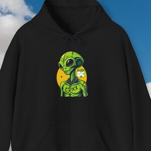 May include: Black hooded sweatshirt featuring a green alien graphic with the text "OG" and "CDXX" on a yellow circle. The alien has large eyes and a detailed design. The sweatshirt has a front pocket and a drawstring hood.