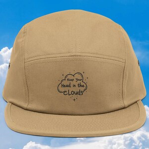 May include: A tan five-panel cap with a flat brim. The cap features the phrase "Keep Your Head in the Clouds" in black script and a cloud graphic. The background is a blue sky with white clouds.