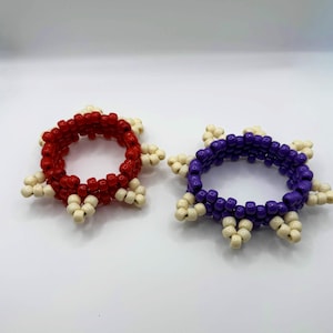 May include: Two beaded bracelets. One is red with cream accents, and the other is purple with cream accents. Each bracelet is circular with a starburst design around the edges, made of small beads.