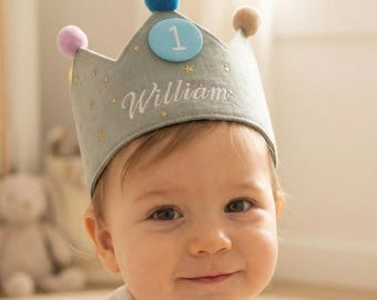 May include: A baby wearing a light gray crown with the name "William" embroidered on it. The crown has a blue circle with the number "1" and is decorated with gold stars and colorful pom-poms. The baby is wearing a white onesie.