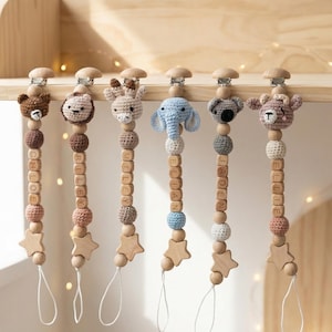 May include: Six baby pacifier clips with crochet animal heads: a bear, lion, giraffe, elephant, koala, and deer. Each clip has wooden beads, a star charm, and a name spelled out in wooden letters. The clips are displayed on a wooden shelf, with a neutral background.