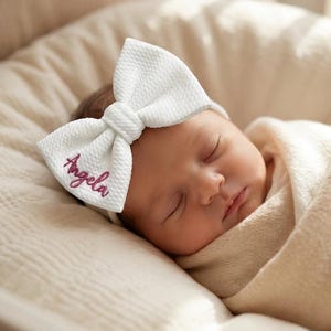 Peut inclure: Un bandeau à nœud en tissu blanc texturé avec le prénom Angela brodé en écriture cursive rose foncé. Le nœud est fixé sur un fin élastique blanc assorti, formant un accessoire décoratif pour la tête avec un détail de nœud visible.