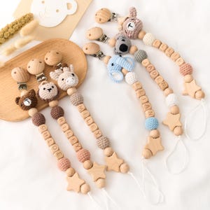 Op de afbeelding: Een verzameling fopspeenclips voor baby's met houten kralen en gehaakte dierenkoppen. Elke clip heeft een ander dier, waaronder een beer, een koala, een giraf en een olifant. De clips hebben namen die in houten letterkralen zijn gespeld.