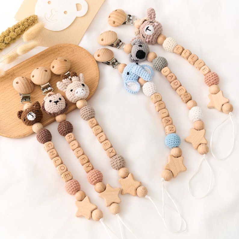 Op de afbeelding: Een verzameling fopspeenclips voor baby's met houten kralen en gehaakte dierenkoppen. Elke clip heeft een ander dier, waaronder een beer, een koala, een giraf en een olifant. De clips hebben namen die in houten letterkralen zijn gespeld.