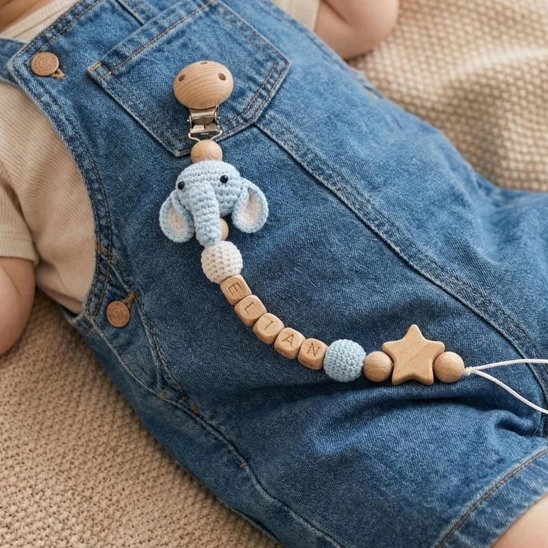 Op de afbeelding: Een baby fopspeenclip met een lichtblauw olifantenontwerp. De clip heeft houten kralen met een naam, een houten ster en een houten clip. De fopspeenclip is bevestigd aan een spijkerbroek.