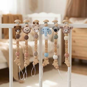 Op de afbeelding: Zes gepersonaliseerde fopspeenclips voor baby's die aan een witte spijlen van een wieg hangen. Elke clip heeft een ander gehaakt dierenhoofd, houten kralen en een houten ster. De namen Chloe, Riley, Miley, Elian, Jaden en Ellie staan op de clips.