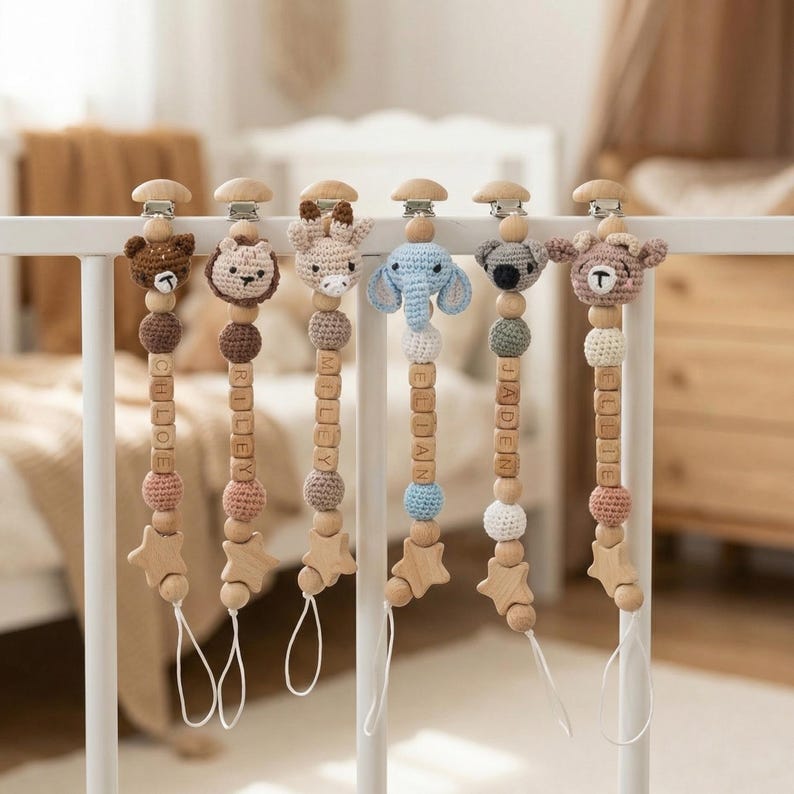 Op de afbeelding: Zes gepersonaliseerde fopspeenclips voor baby's die aan een witte spijlen van een wieg hangen. Elke clip heeft een ander gehaakt dierenhoofd, houten kralen en een houten ster. De namen Chloe, Riley, Miley, Elian, Jaden en Ellie staan op de clips.