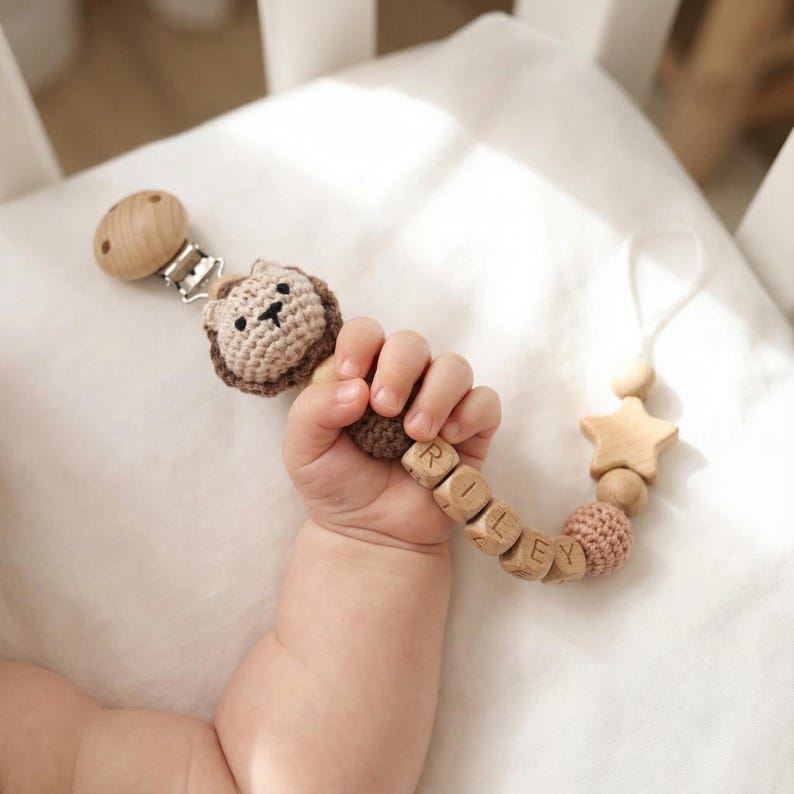 Op de afbeelding: Een houten fopspeenclip, vastgehouden door de hand van een baby. De clip heeft een gehaakte leeuwenkop, houten kralen met de tekst "RILEY", een stervormige kraal en een houten clip. De fopspeenclip is lichtbruin en beige.
