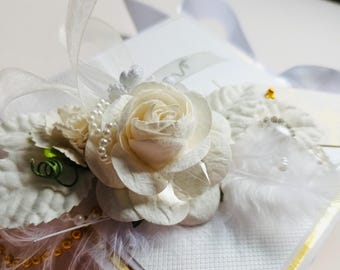 Puede incluir: Un arreglo floral blanco con una gran rosa de papel, perlas y plumas. El arreglo está sobre una caja blanca con ribete dorado. Una cinta blanca y otros elementos decorativos completan el diseño.