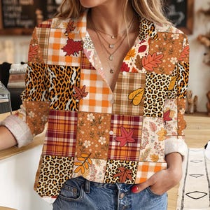 May include: A long-sleeve button-down shirt featuring a patchwork design of orange and brown plaid, leopard print, and autumn leaf patterns. The shirt has a collared neckline and is worn tucked into blue denim jeans, showcasing a layered print style.
