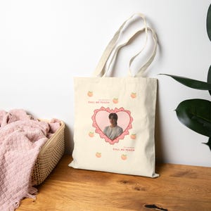 May include: Cream-colored tote bag featuring a heart-shaped design with a person's image inside. The heart is outlined with a pink lace design. Peaches and the text "CALL ME PEACH" are scattered across the bag.